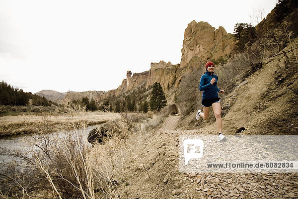 A athletic woman in a red hat jogs down a trail leading along a riverside in Smith Rock Oregon.