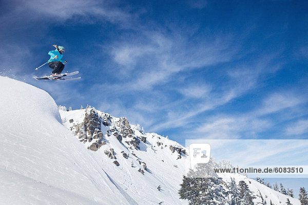A female skier launching into fresh powder snow with mountains and blue sky in the background.