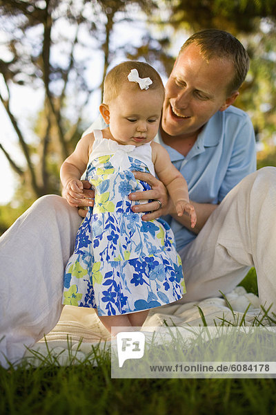 A father plays with his baby girl  while sitting on a picnic blanket outside on a sunny day in California.