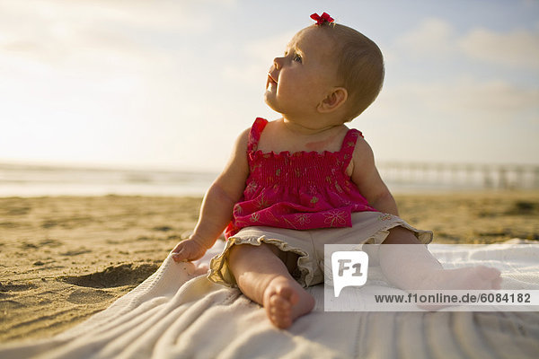 A baby girl with a red bow in her hair looks up to the sky while sitting on a blanket in the sand during a sunny day at the beach in California.