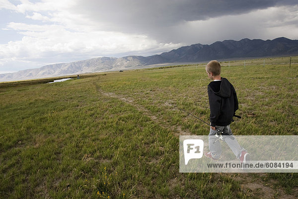A boy walking with his fishing gear through a field  with storm clouds in the distance.