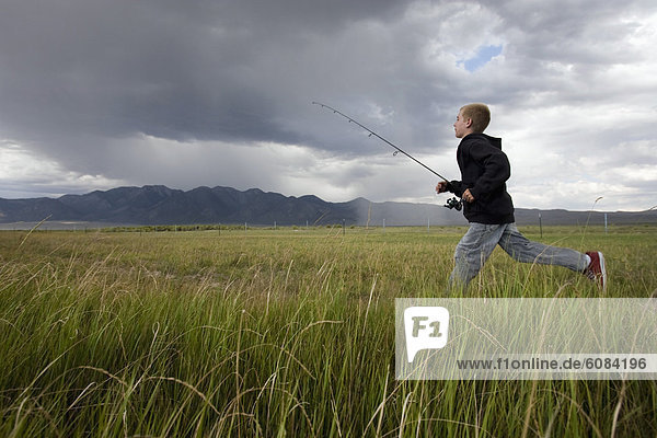 A boy runs with his fishing gear through a field  with storm clouds in the distance.