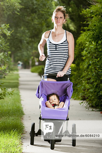 An edgy-looking  athletic young woman enjoys a run on a suburban sidewalk with a baby in a jogging stroller.