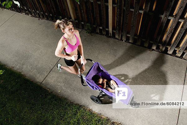 An edgy-looking  athletic young woman in a sports bra enjoys a run on a suburban sidewalk with a baby in a jogging stroller.