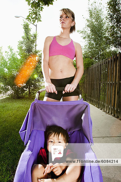 An edgy-looking  athletic young woman with tattoos stops during a run on a suburban sidewalk with a baby in a jogging stroller.