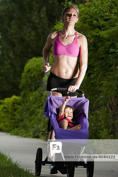 A baby raises his fist and smiles in his jogging stroller while being pushed by an edgy-looking  athletic young woman during a run on a suburban sidewalk.