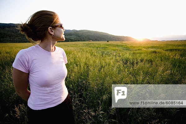 A girl in sunglasses walks in the tall grass watching the sunset.