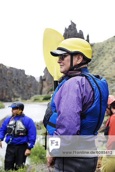 An older man outfitted in river gear stands on the Owyhee River bank  holding a paddle and waiting for instruction.