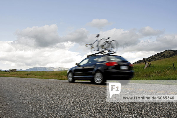 A black car with two road bikes on top speeds down a country road just outside Bozeman Montana. The Bridger Mountain range can be seen in the distance.