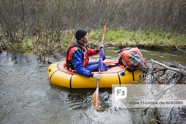 A young man paddles his small  inflatable raft through a thicket of trees.