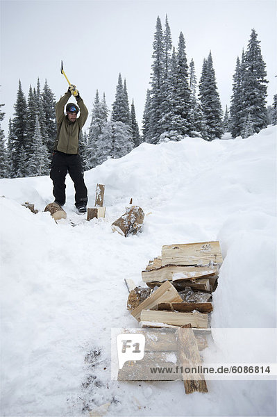 A man chops wood in his snowboarding gear in the snowy backcountry.