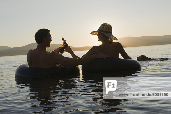A young couple relax with a cold beverage while floating in a lake at sunset in inner tubes.