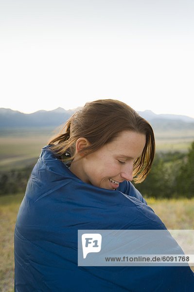 A young woman wraps herself in a sleeping bag as the sun sets.