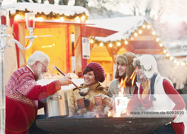 Österreich  Salzburg  Senior Mann  der jungen Leuten auf dem Weihnachtsmarkt einen Drink serviert  lächelnd