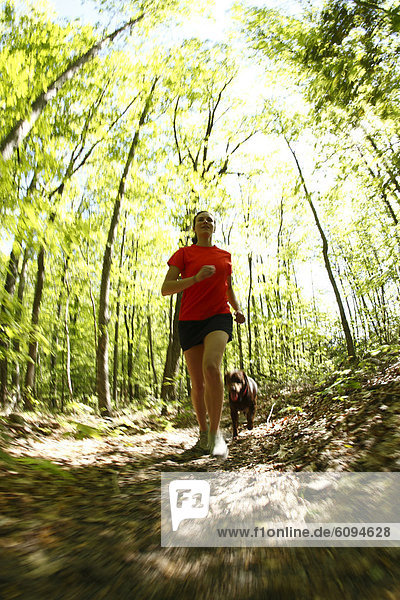Woman trail running in a lush green forest with her dog.