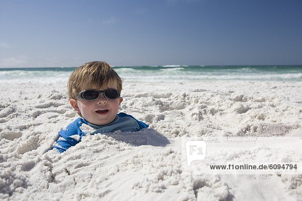 A little boy with sunglasses is buried in the sand up to his head with blue sky and ocean in the background.