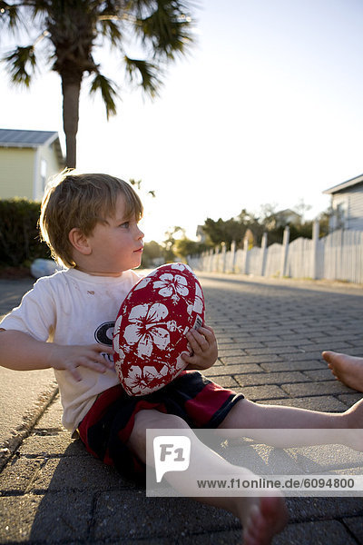 A little boy is holding a football in an alley with palm trees and sun in the background.