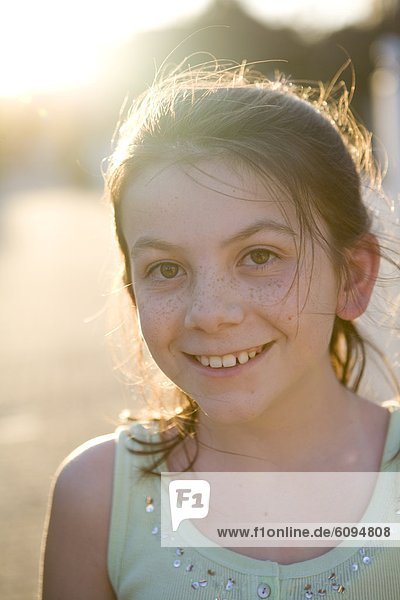 A little girl with freckles is smiling at the camera with the sun shining in the background.