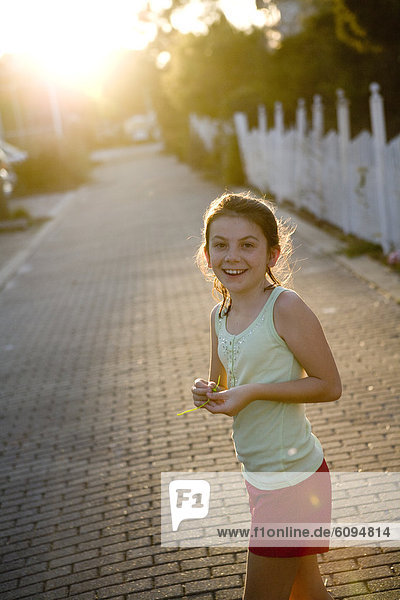 A little girl wearing shorts is smiling at the camera with a picket fence and sun in the background.