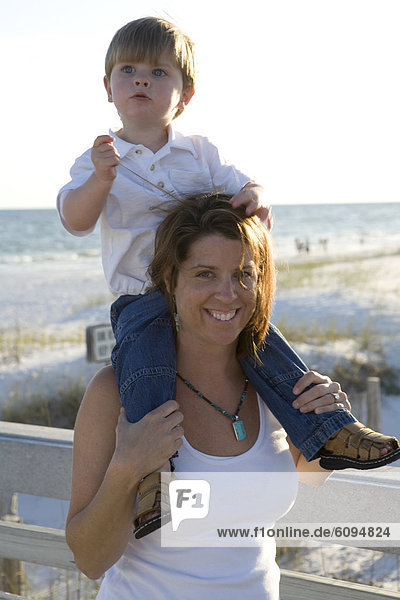 A mom is holding a little boy on her shoulders smiling with the beach and ocean in the background.
