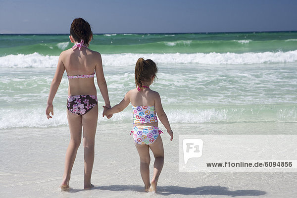 Two little girls in bikinis are holding hands at shoreline with the ocean in the background.