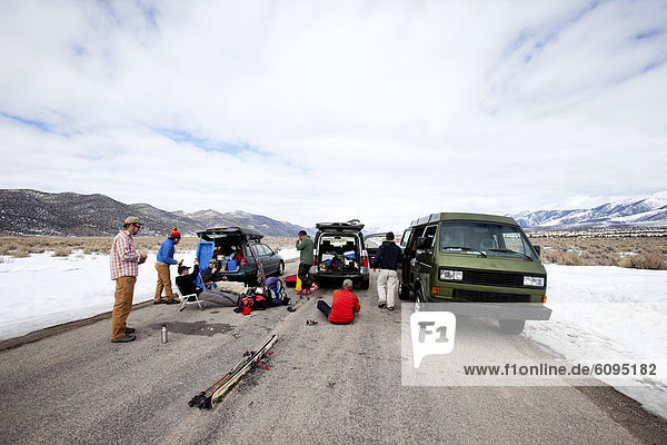 Six men hanging out in the middle of a road with their ski gear and vehicles in a remote mountainous area.