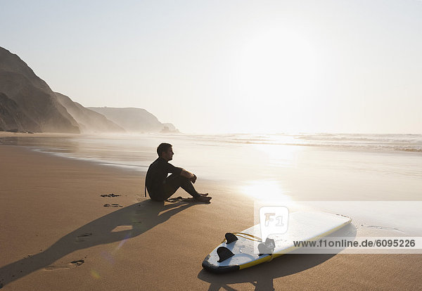 Portugal  Junger Mann am Strand am Surfbrett sitzend