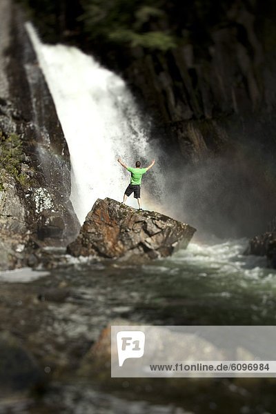 A young man holds his arms open standing at the base of a waterfall after rappeling down.