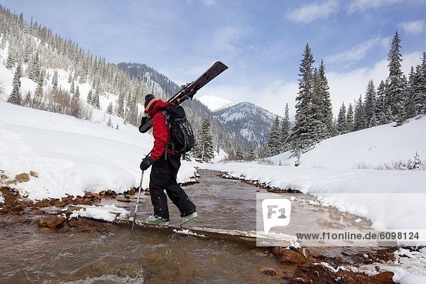 A man carries his skis across a snowy  mountain stream.