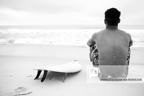 Male surfer sits by surfboard at the beach and looks at the ocean.
