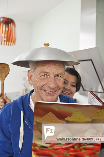 Senior man holding cook book with wok lid on head  woman holding spatula in background