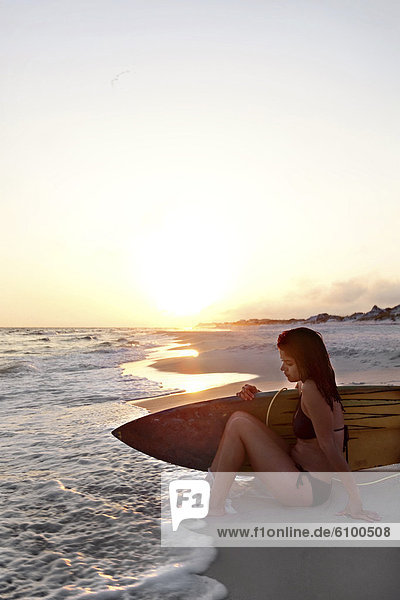 young woman sitting on beach in bikini with surfboard. backlit sunset with surf coming up to her feet.