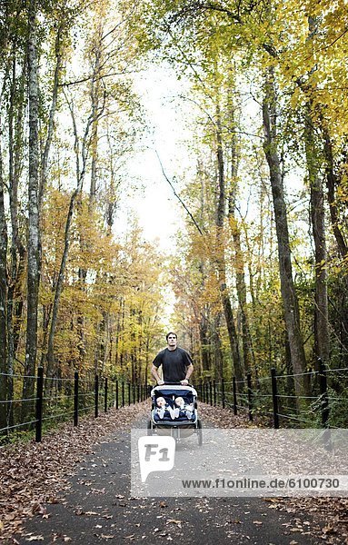 A man runs on a greenway in the fall with his twin sons in a stroller.