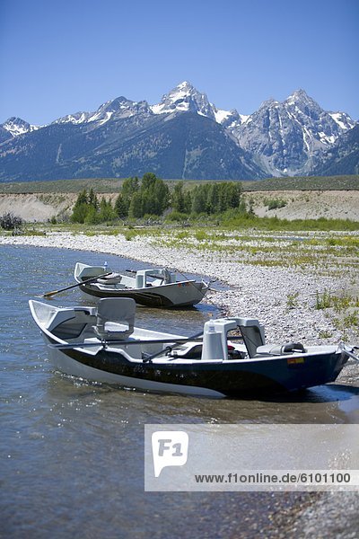 Two river row boats sit on the shore of the Snake River with snow capped mountains in the background.