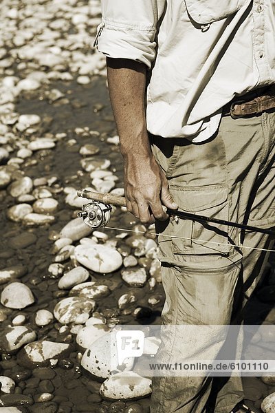 A fisherman holds his fly fishing rod on his side with white river rocks behind him.