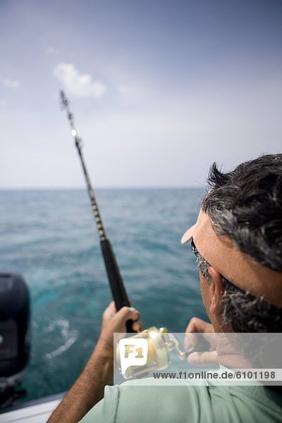 An over the shoulder view of a fisherman reeling in fish with blue water in the distance.
