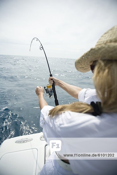 A blonde woman in a straw hat reels in a fish with blue water in the distance.