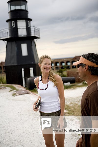 A young blonde woman in shorts looks at a man and smiles with an old black lighthouse in the background.
