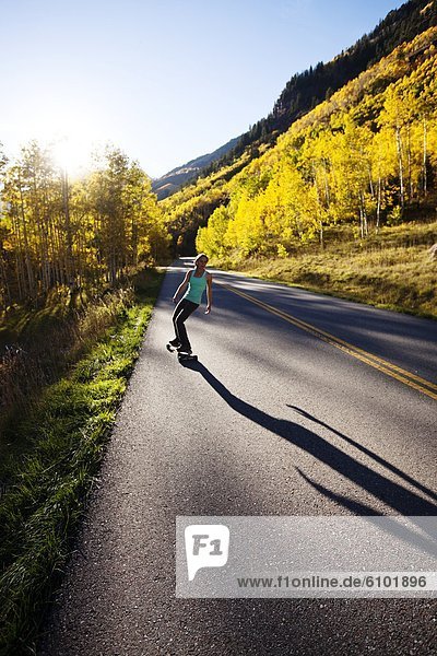 A young woman longboards down a smooth country road through the mountain peaks and gold forests.