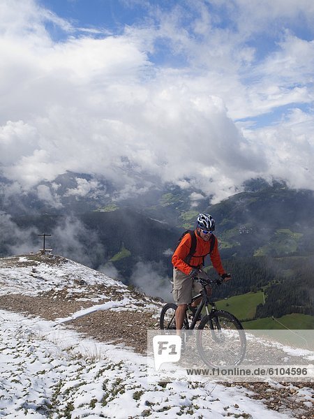 A mountain biker arrives on the summit of Kronplatz  mountains  a cross and clouds in the background.