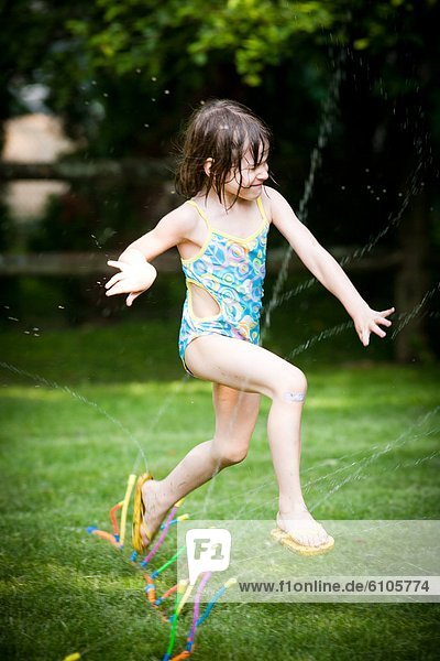 A five-year-old girl jumps through a sprinkler in her bathing suit in her back yard.