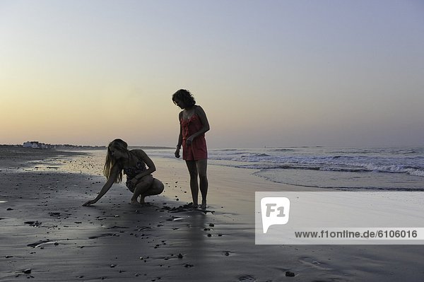 Two young women search for shells at sunset on the beach.