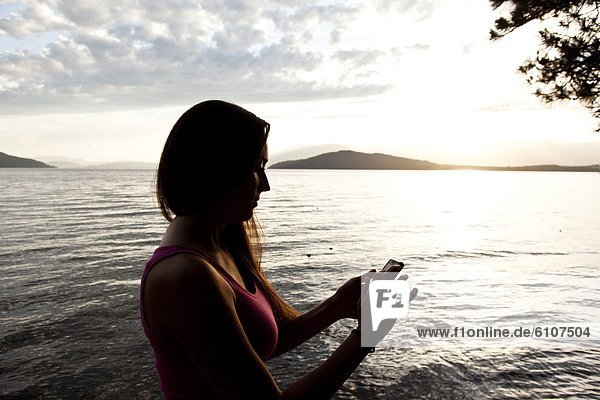 A beautiful young woman playing on her smart cell phone at sunset next to a lake in Idaho.
