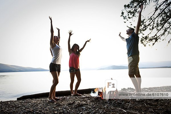 Three young adults smiling and dancing around a camp fire on a camping and kayaking trip on a lake in Idaho.