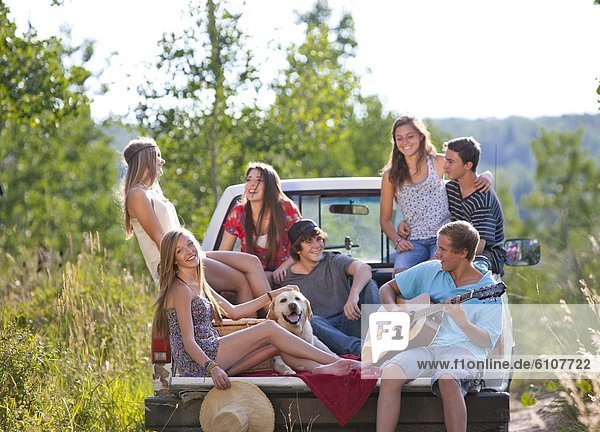 A group of teen-aged friends enjoy a SUmmer afternoon laughing and singing in the back of a truck along a country road.