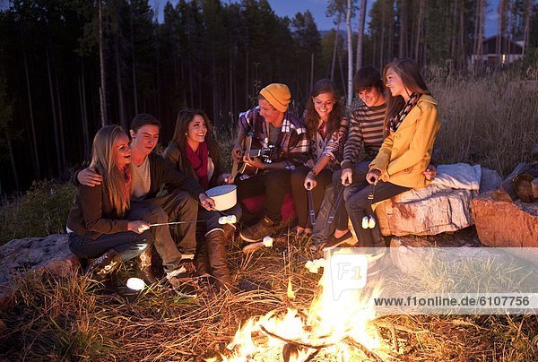 A group of teen-aged girls and boys sing and roast marshmallows around a camp fire on a Summer evening.