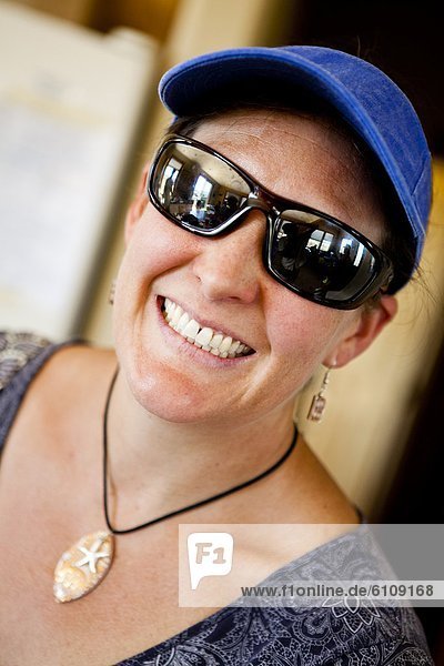 A woman wears sunglasses  visor and a seashell necklace while she smiles at the camera.