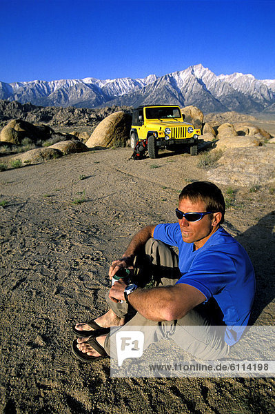 Man with sunglasses sitting in the desert next to his jeep in front of mountains.