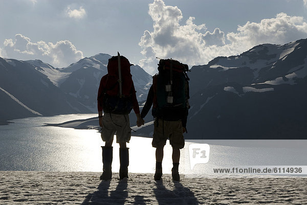 Two backpackers hold hands while overlooking a high alpine lake and distant mountains.