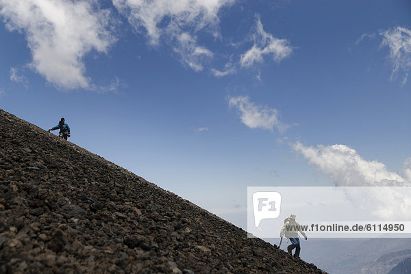 Two climbers descend a steep scree slope with a blue sky in the background.